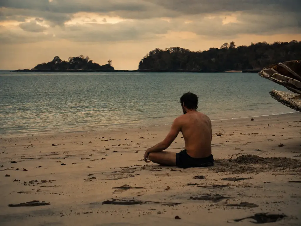 Man meditates on a beach with a jungle in the background.