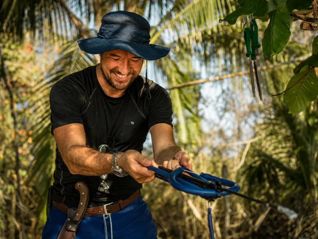 Man in hat smiles while preparing speargun in tropical setting.